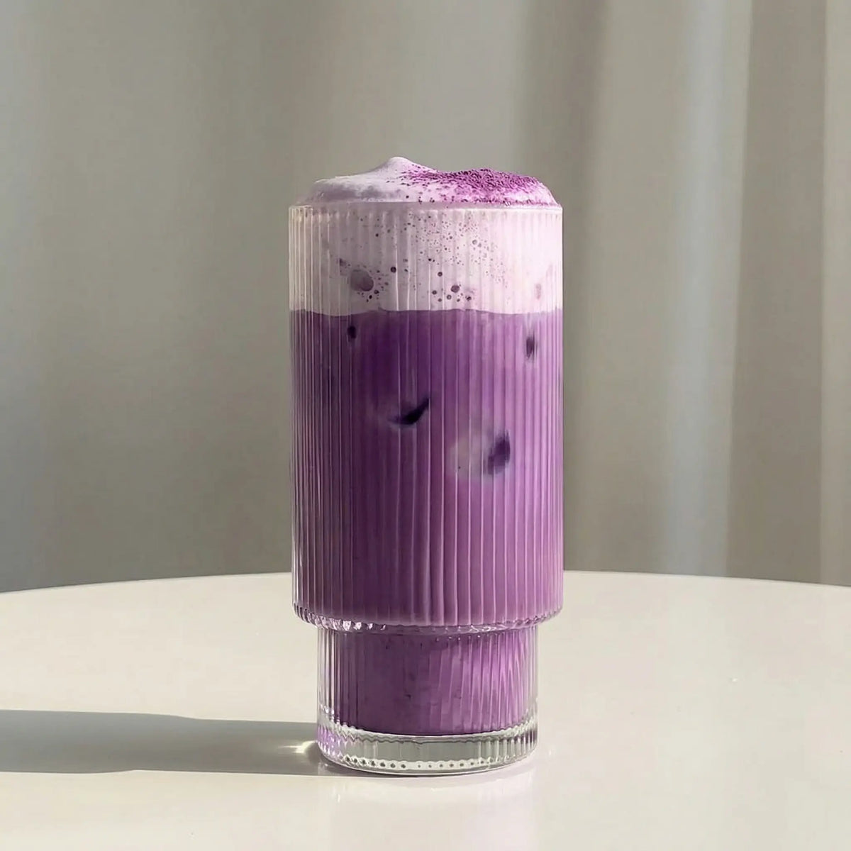 Ube latte violet intense à la poudre d'ube dans un verre cannelé, surmonté d'une mousse crémeuse saupoudrée d'ube
